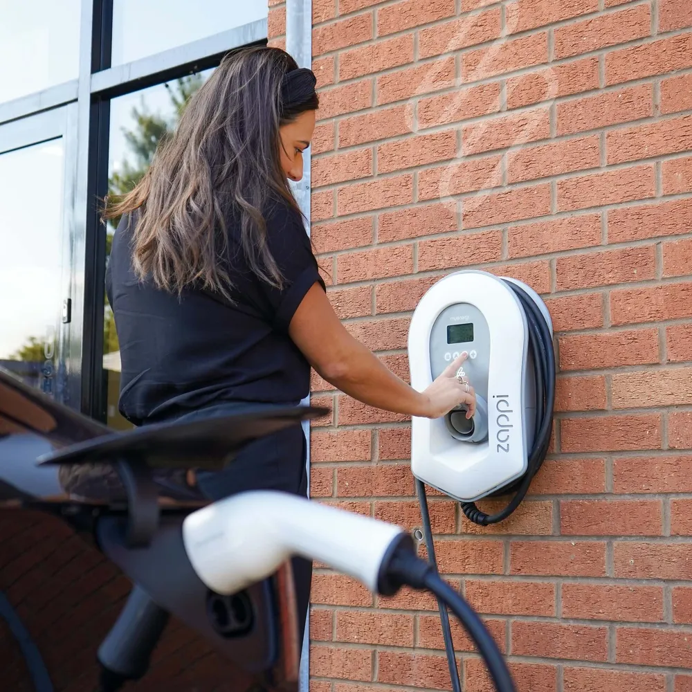 A driver adjusting a wall-mounted EV charger beside her vehicle